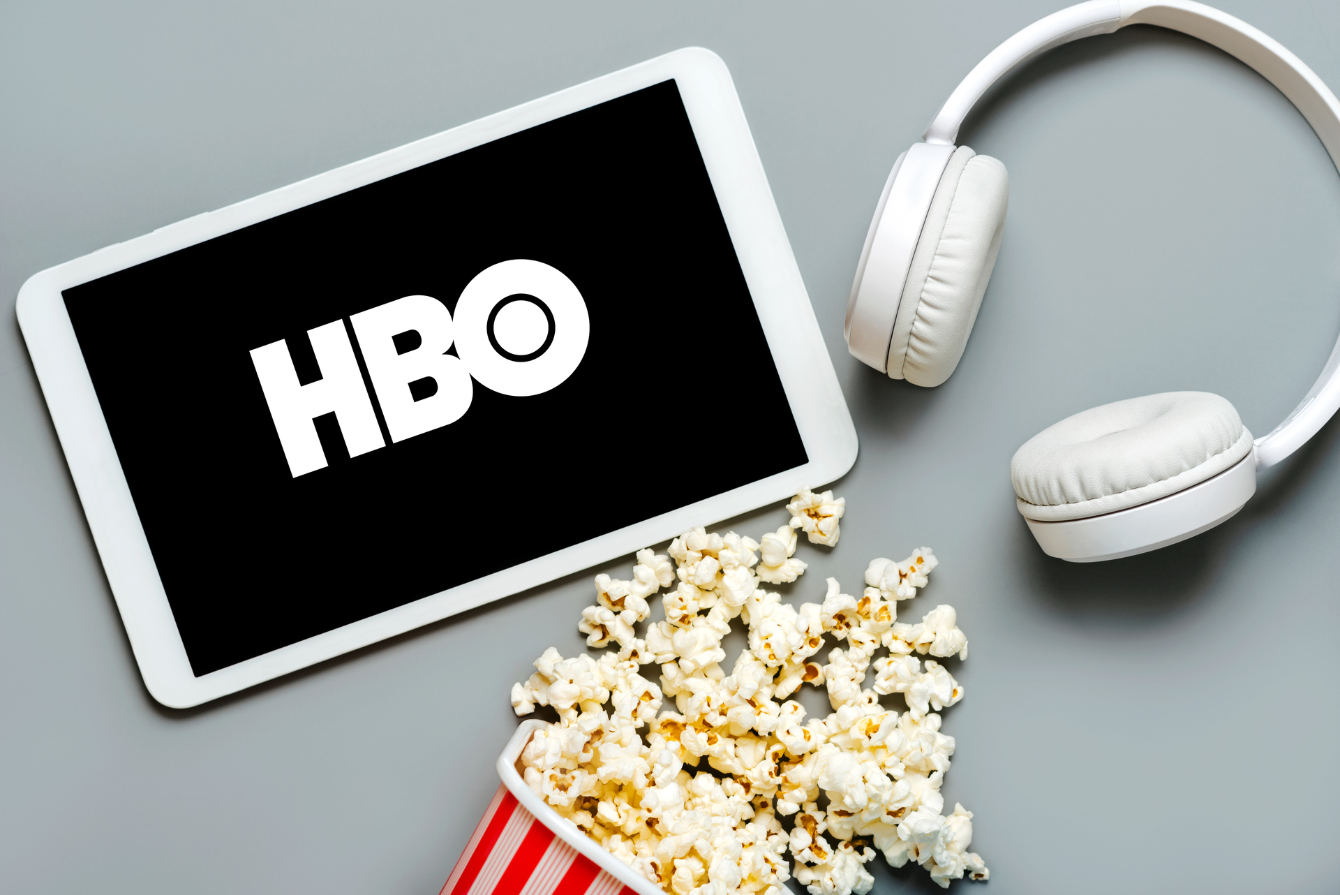 Fotografia zobrazuje tablet s logom HBO na obrazovke, biele slúchadlá vedľa tabletu a červeno-bielu pruhovanú nádobu popcornu, ktorá sa rozsypala vedľa nich. Farba pozadia je tlmená sivá