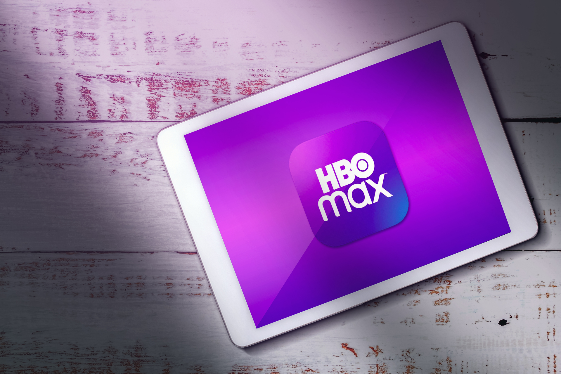 Na obrázku je zobrazený tablet alebo digitálne zariadenie so žiarivou fialovou obrazovkou, na ktorej je nápadne zobrazené logo „HBO Max“. Tableta spočíva na drevenom povrchu so starým alebo ošúchaným vzhľadom, ktorý má biely povrch s presvitajúcimi škvrnami s červeno-ružovými podtónmi. Celkové osvetlenie dodáva obrazu jemnú a náladovú atmosféru