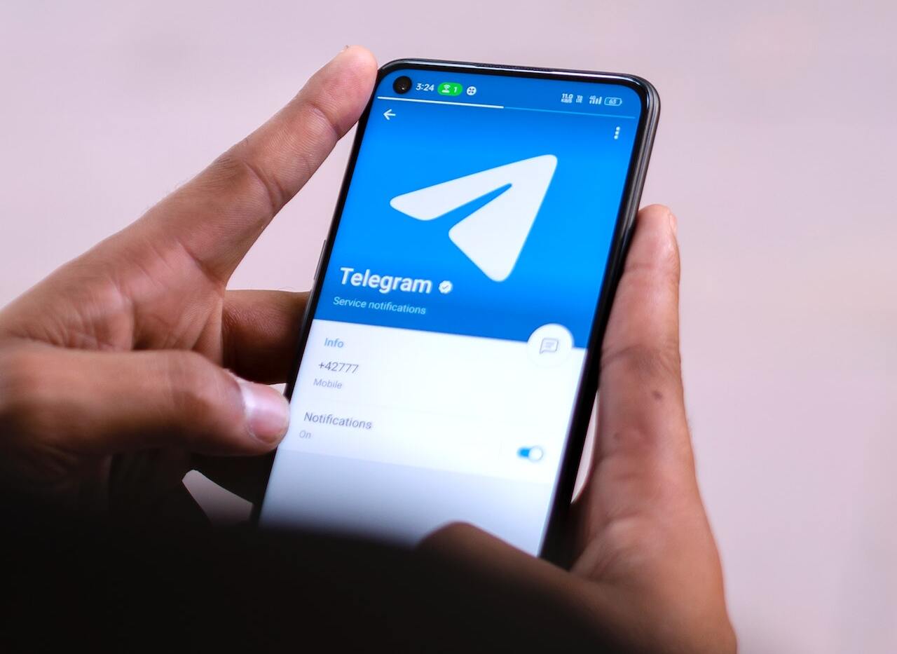 Na fotografii môžeme vidieť ruku držiacu mobil s otvorenou aplikáciou Telegram. Na obrazovke mobilu je zobrazené logo Telegramu a niekoľko možností nastavenia, vrátane notifikácií